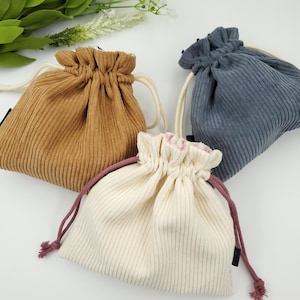 May include: Three drawstring bags made of corduroy fabric. The bags are in beige, white, and gray.