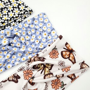 May include: Three twisted headbands featuring fabric patterns: one black with white and yellow daisies, one light blue with white and yellow daisies, and one white with orange flowers and brown and yellow butterflies.