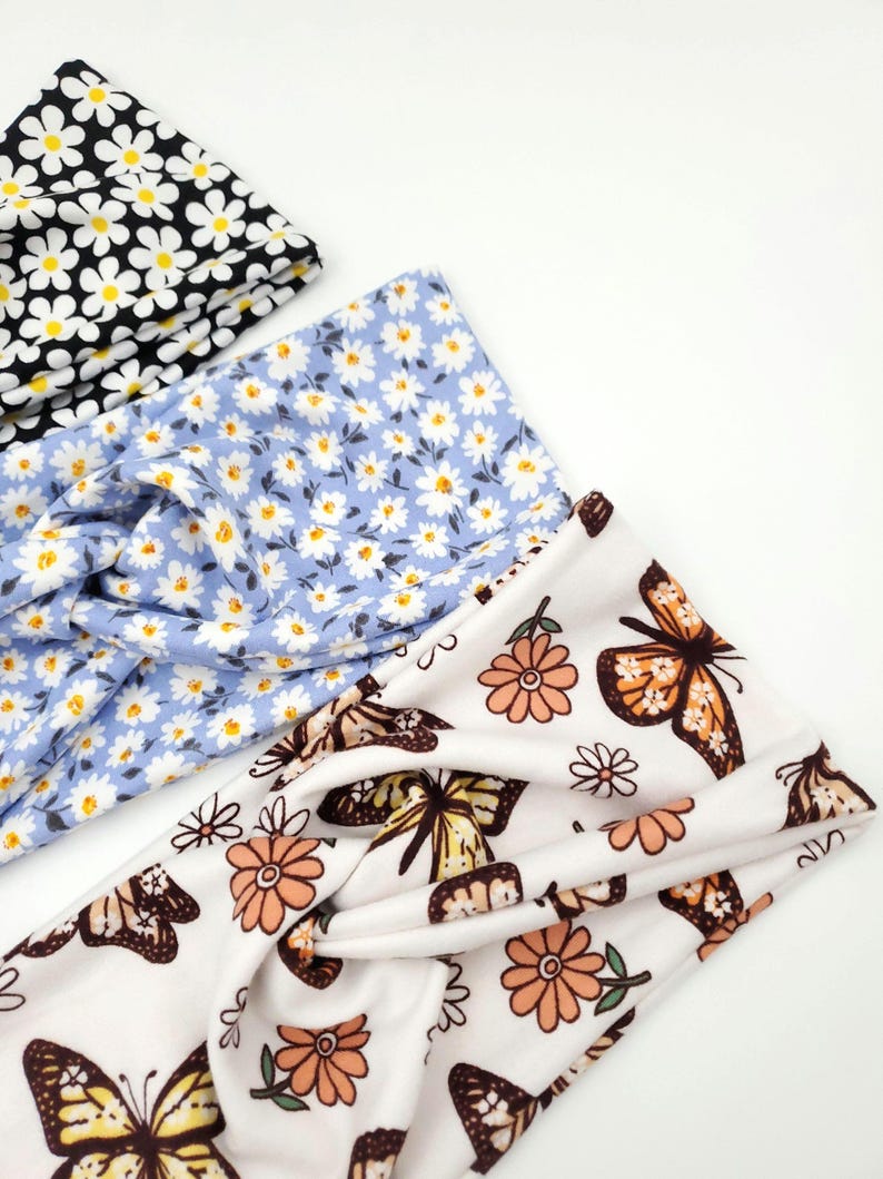 May include: Three twisted headbands featuring fabric patterns: one black with white and yellow daisies, one light blue with white and yellow daisies, and one white with orange flowers and brown and yellow butterflies.