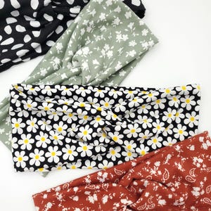 May include: Four fabric headbands with twisted front details. The top headband is black with white spots. The second is sage green with white flowers. The third is black with white daisies and yellow centers. The bottom is rust orange with white floral patterns.