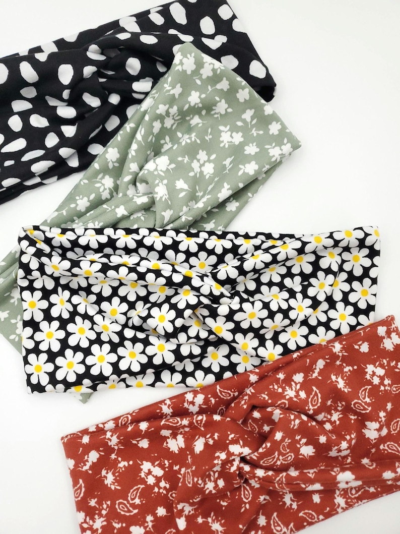 May include: Four fabric headbands with twisted front details. The top headband is black with white spots. The second is sage green with white flowers. The third is black with white daisies and yellow centers. The bottom is rust orange with white floral patterns.