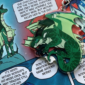 Op de afbeelding: Een groene draak met een lichtgroene buik en donkergroene vlekken is afgebeeld in cartoon-stijl. De draak zit op een rots met zijn vleugels uitgespreid. De draak heeft een woeste uitdrukking op zijn gezicht. De afbeelding is een sleutelhanger.