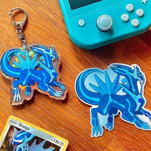 Könnte beinhalten: Zwei blaue und weiße Schlüsselanhänger mit Pokémon-Charakteren. Einer ist ein Schlüsselanhänger mit transparentem Hintergrund, der andere ein Aufkleber. Beide zeigen das Pokémon Dialga.