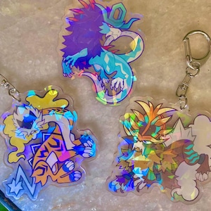 Può includere: Tre portachiavi olografici iridescenti con diverse creature mitiche. Il primo portachiavi presenta un drago blu e viola con una gemma verde sulla testa. Il secondo portachiavi presenta una tigre gialla e arancione con un fulmine. Il terzo portachiavi presenta un lupo marrone e arancione con una criniera gialla.