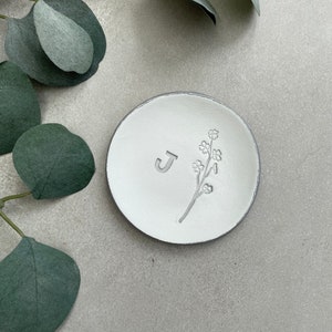 May include: White ceramic ring dish with a gray rim. The dish has a small floral design and the letter "J" embossed on the surface.