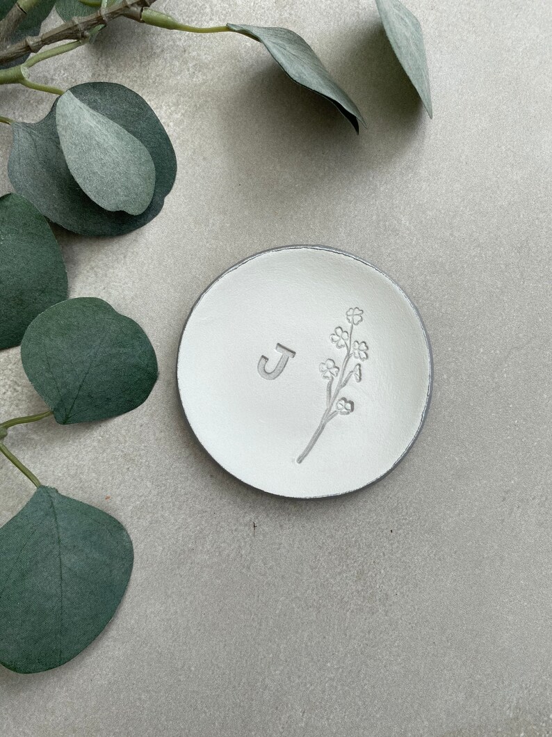 May include: White ceramic ring dish with a gray rim. The dish has a small floral design and the letter "J" embossed on the surface.