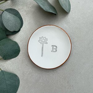May include: White ceramic ring dish with a brown rim. The dish has a floral design and the letter "B" engraved on it.