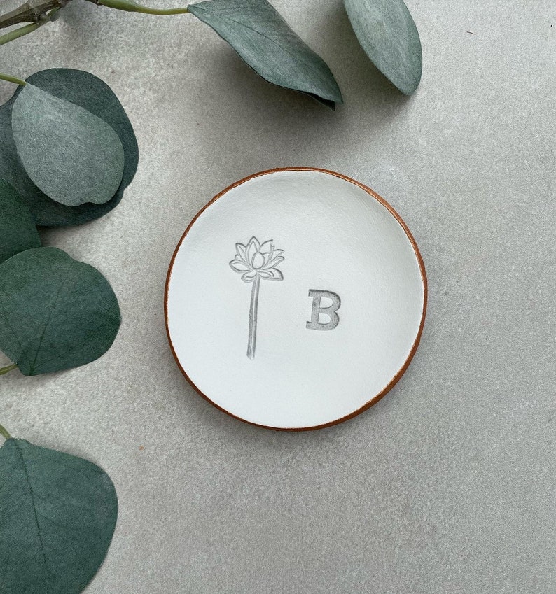 May include: White ceramic ring dish with a brown rim. The dish has a floral design and the letter "B" engraved on it.