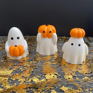 May include: Three white ghost figurines with black dot eyes and orange pumpkin accents. One ghost holds a pumpkin, while the others wear pumpkin hats. The Halloween-themed decorations are set against a black background.