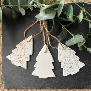 May include: Three white ceramic Christmas tree decorations with twine hangers. Two trees have gold and silver speckled designs. The decorations are displayed on a black surface with green foliage.