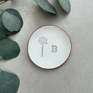 May include: White ceramic ring dish with a brown rim. The dish has a floral design and the letter "B" embossed on the surface.