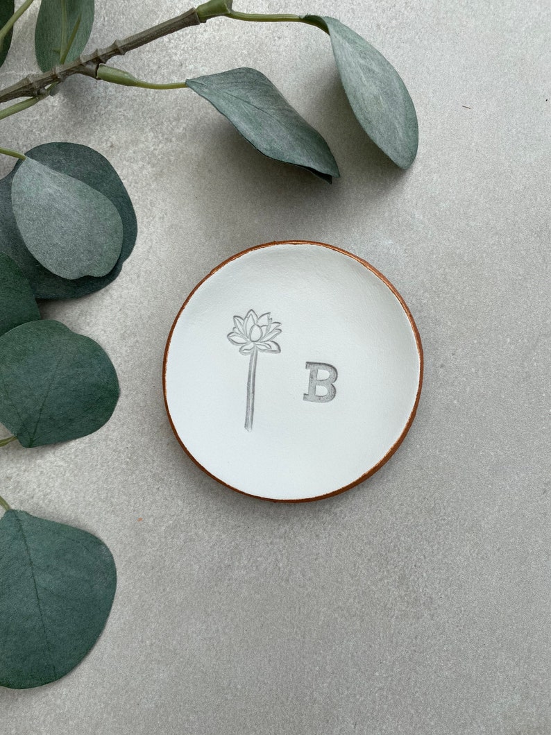 May include: White ceramic ring dish with a brown rim. The dish has a floral design and the letter "B" embossed on the surface.