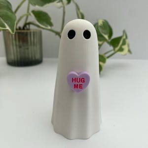 May include: A white ghost figurine with two black circular eyes. The ghost has a light purple heart-shaped accent on its chest with the text HUG ME printed in red capital letters. The figurine has a smooth, matte finish and a rounded top.
