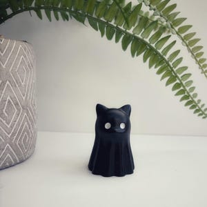 May include: A small, solid black figurine shaped like a cat ghost. The object features a smooth, matte finish with two distinct, circular white eyes positioned on the front. The base is wide and slightly flared, tapering upward to pointed ears.
