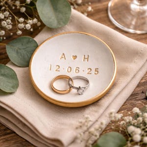 May include: A white ceramic ring dish with a gold rim, holding two rings. The dish is inscribed with "A ♡ H" and "12.06.25". The rings include a gold band and a silver ring with a diamond. The dish rests on a folded beige linen napkin.