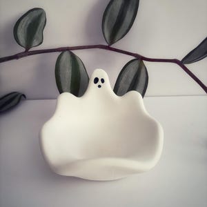 May include: A white ceramic dish shaped like a ghost with a smooth, matte finish. The ghost figure features a small, hand-painted face consisting of two oval eyes and a circular mouth in black, positioned at the top centre of the curved rim.