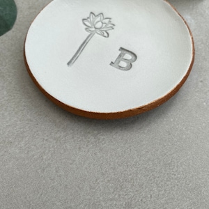 May include: White ceramic ring dish with a gold rim. The dish has a small flower design and the letter "B" embossed on the surface.