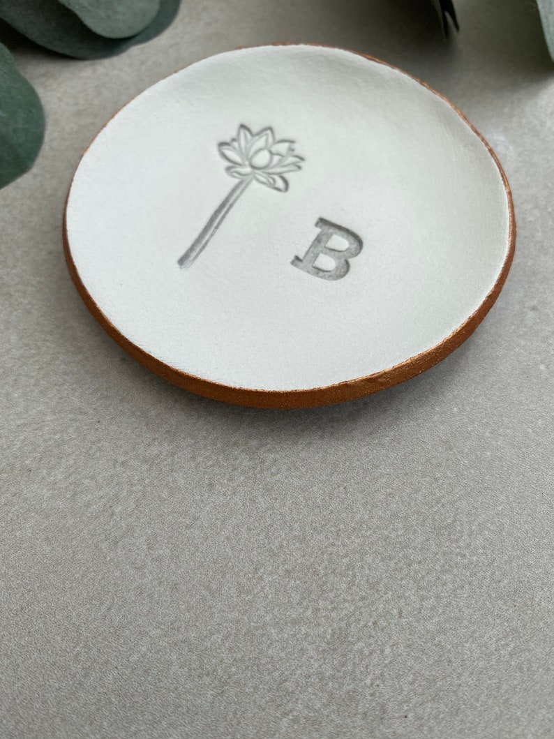 May include: White ceramic ring dish with a gold rim. The dish has a small flower design and the letter "B" embossed on the surface.