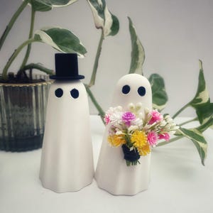 May include: Two white ghost figurines with black eyes. One ghost wears a small black top hat. The other ghost holds a small bouquet of colourful pink, yellow, and white artificial flowers wrapped in black string. The figures have a smooth finish.
