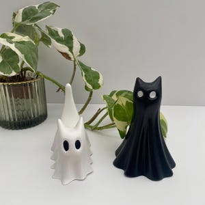 May include: Two Halloween-themed figurines. One is a white ghost with black oval eyes. The other is a black cat-shaped figure with white eyes. Both are standing on a white surface, with a potted plant in the background.