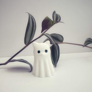May include: A small, matte white figurine shaped like a ghost with pointed ears and two round, dark eyes. The ghost has a smooth, draped body with vertical folds at the base, standing upright as a decorative object with a simple, clean aesthetic.