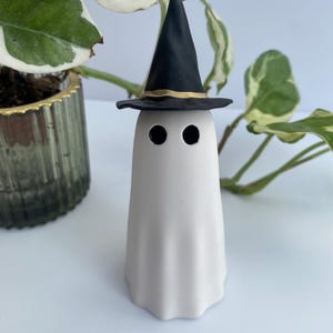 May include: A white ghost figure wearing a black witch's hat with a gold trim. The ghost has two black eyes and a draped form. A Halloween decoration.