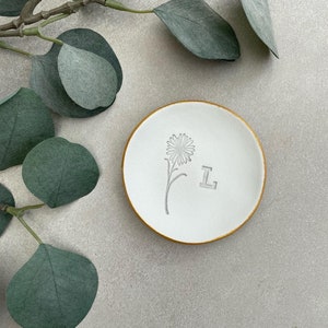 May include: White ceramic trinket dish with a gold rim. The dish has a small flower and the letter "L" embossed on the surface.
