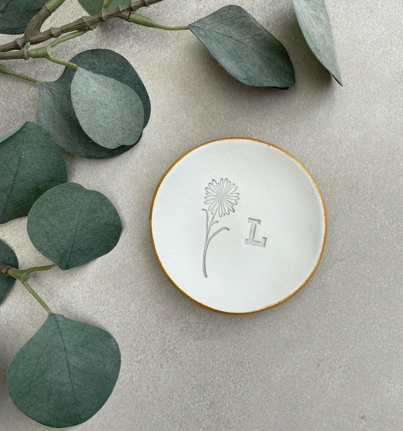 May include: White ceramic trinket dish with a gold rim. The dish has a small flower and the letter "L" embossed on the surface.