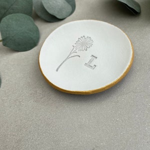 May include: White ceramic ring dish with a gold rim. The dish has a simple flower design and the letter "L" embossed on the surface.