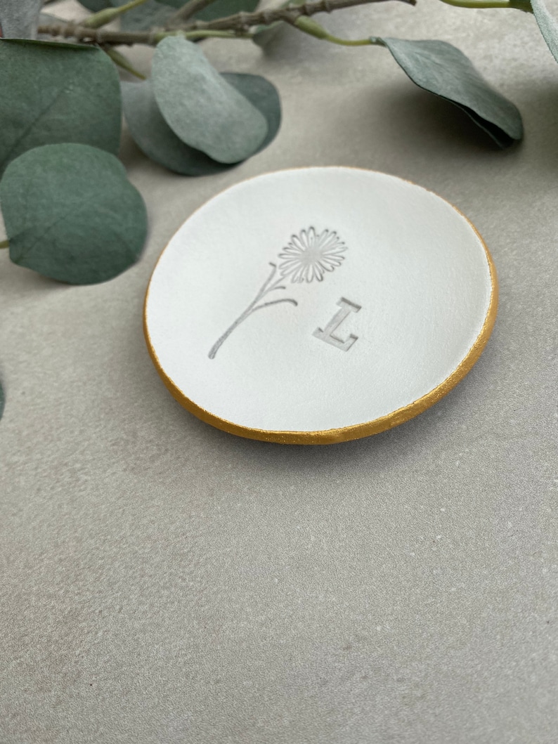 May include: White ceramic ring dish with a gold rim. The dish has a simple flower design and the letter "L" embossed on the surface.