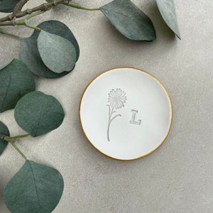 May include: White ceramic ring dish with a gold rim. The dish has a simple flower design and the letter "L" embossed on the surface.