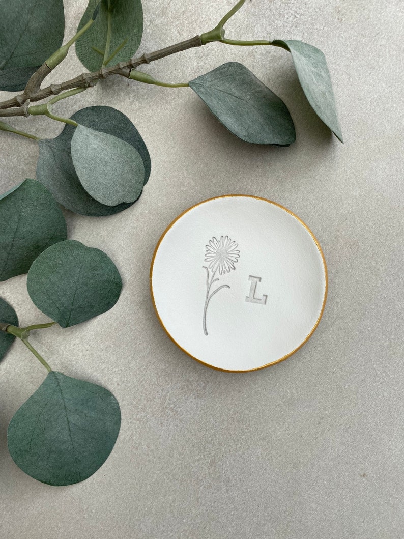 May include: White ceramic ring dish with a gold rim. The dish has a simple flower design and the letter "L" embossed on the surface.