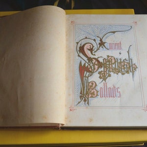 May include: Open book with the title "Ancient Scottish Ballads" in ornate calligraphy. The page features a decorative border with red, gold, and blue accents. The book is open on a yellow surface.