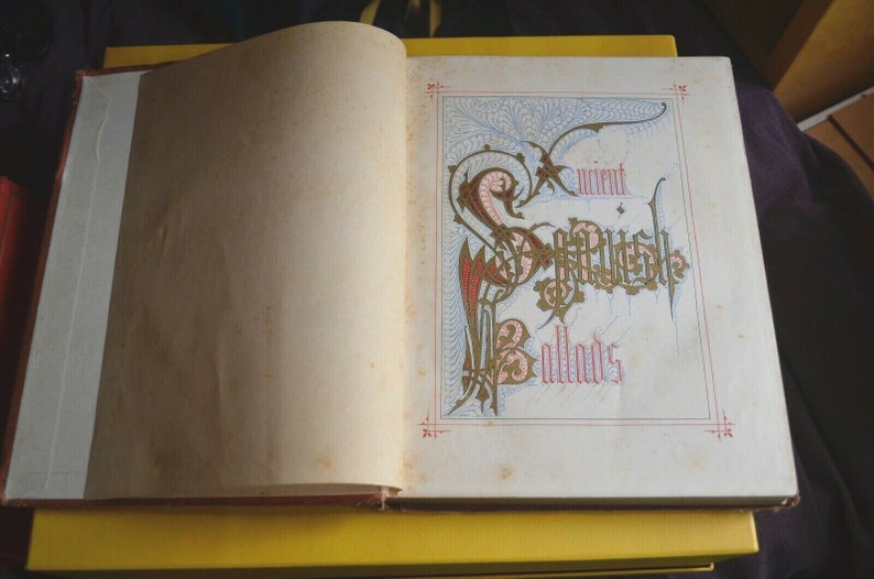 May include: Open book with the title "Ancient Scottish Ballads" in ornate calligraphy. The page features a decorative border with red, gold, and blue accents. The book is open on a yellow surface.