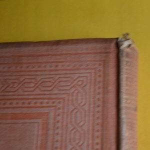 May include: Close-up of a vintage book's corner, showcasing a detailed, embossed design in a reddish-brown hue. The cover's texture and intricate patterns are visible, with a yellow background.