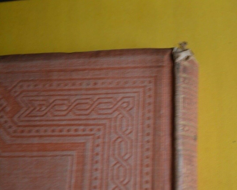 May include: Close-up of a vintage book's corner, showcasing a detailed, embossed design in a reddish-brown hue. The cover's texture and intricate patterns are visible, with a yellow background.