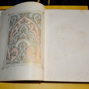 May include: Open book with an ornate, colorful illustration on the left page. The design features pink, blue, and gold accents on a white background. The right page is blank with a slightly aged appearance. The book is open on a yellow surface.