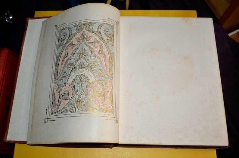 May include: Open book with an ornate, colorful illustration on the left page. The design features pink, blue, and gold accents on a white background. The right page is blank with a slightly aged appearance. The book is open on a yellow surface.