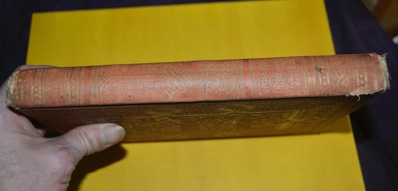 May include: Side view of an antique book with a worn, reddish-brown cover. The spine features embossed gold geometric patterns and text. The book's edges show signs of age and wear, suggesting it is a vintage item.