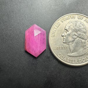May include: A faceted, hexagonal pink gemstone positioned next to a United States quarter coin for scale. The gemstone displays a vibrant magenta hue with visible internal facets and a pointed, elongated shape, contrasting against the silver coin.