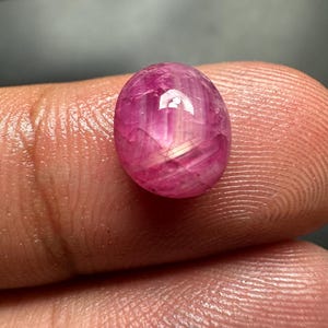 Puede incluir: Una gema ovalada pulida de un tono rosa vibrante. La gema tiene una superficie lisa con reflejos internos que crean un brillo sutil. La piedra se sostiene entre los dedos, mostrando su tamaño y color.