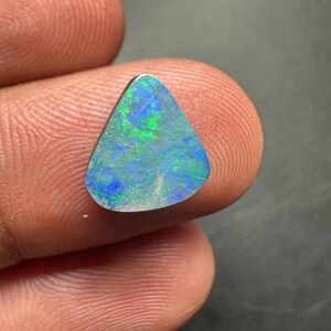 May include: A triangular-shaped opal gemstone with a vibrant display of colors. The opal features a mix of blue, green, and hints of yellow, creating a shimmering effect. The stone is held between two fingers, showcasing its size and shape.