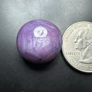 May include: A polished, round, purple gemstone with a white inclusion, next to a U.S. quarter for size comparison. The gemstone has a unique, textured appearance and is set against a dark background.