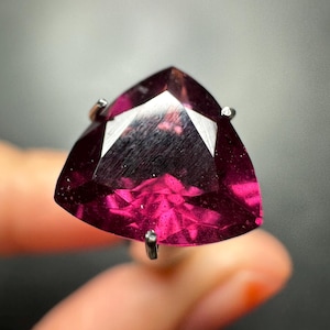 Puede incluir: Una piedra preciosa de corte trillón en color magenta intenso montada en un engaste de garras de metal plateado. La piedra presenta una superficie facetada que refleja la luz, mostrando un tono rosa rojizo vibrante y saturado.