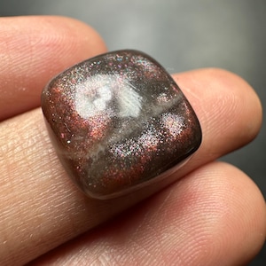 May include: A square-cut gemstone with a dark brown hue, featuring sparkling red and silver inclusions. The stone has a smooth, rounded finish. The gemstone is held in a person's hand. The unique colour and texture of the stone make it a striking piece.