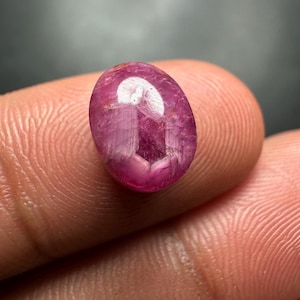 以下が含まれることがあります： 磨かれた楕円形のルビーの宝石。宝石は、内部の光の反射を伴う深いピンクがかった赤色です。ルビーは2本の指の間に保持され、そのサイズと透明度を示しています。宝石は、自然な、未装着の石です。