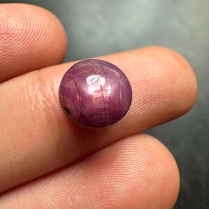 May include: A round, cabochon-cut gemstone with a deep magenta and purple hue. The surface of the stone displays a distinct six-rayed star effect, known as asterism, which is visible under the light reflecting off the polished, smooth surface.