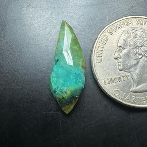 May include: A faceted, teardrop-shaped gemstone featuring a blend of turquoise, green, and yellow hues with dark inclusions. The stone is positioned next to a United States quarter coin for scale, highlighting its relative size and geometric cut.