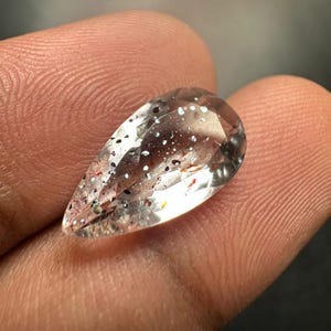 Pode incluir: Uma pedra preciosa transparente lapidada em forma de pera, apresentando inclusões internas de pequenas manchas minerais dispersas em cinza escuro, preto e laranja avermelhado por toda a estrutura cristalina, refletindo a luz na superfície.