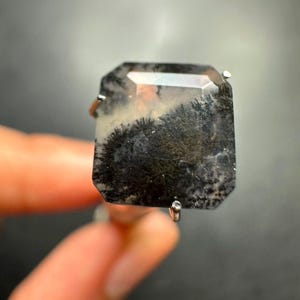 May include: A ring featuring a rectangular, faceted dendritic agate gemstone. The stone is translucent with a milky white base and prominent, dark black, fern-like inclusions branching throughout the interior. It is set in a simple silver-toned metal prong setting.